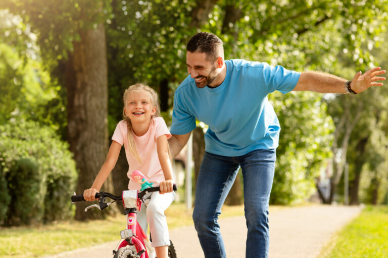 Happy father teaching his child daughter to ride a bike in the park, enjoying time together on weekend on nature.