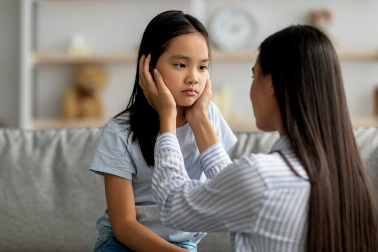 Young mother comforting her sad offended daughter after quarrel at home. Asian woman calming down her child, sitting together in living room interior