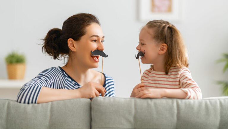 Mom and her child daughter are playing at home. Cute girl are holding paper mustache on stick. Family holiday and togetherness.