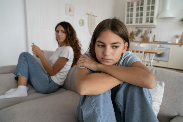 Upset offended teen girl daughter sitting separately with mother on sofa, mom and teenage child ignoring each other after fight at home, selective focus. Generation gap between teenagers and parents
