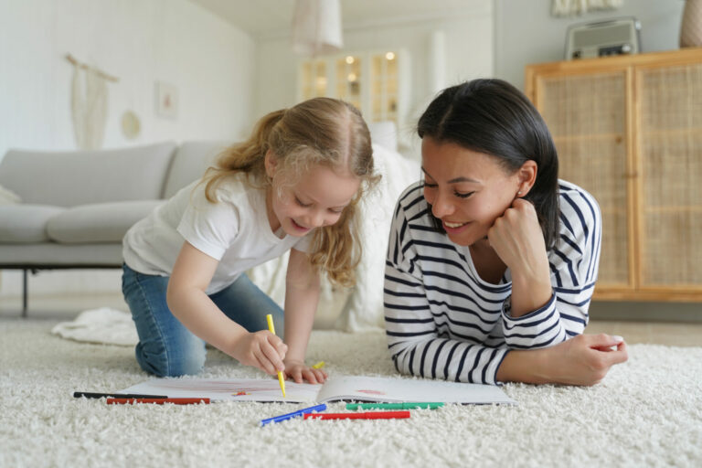 Smiling mother and little adopted child daughter painting together, lying floor. Mom or babysitter helps preschooler kid girl to draw with pencils.