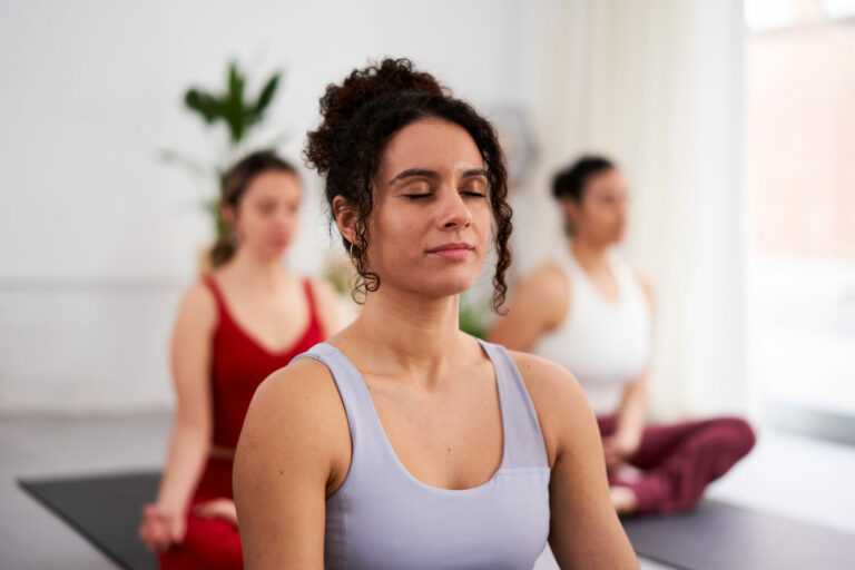 Close up of a young female instructor in yoga lessons, sitting in Sukhasana, meditating inside studio. Front view. Wellness and mental healthcare help.